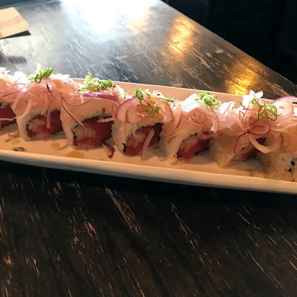 A long white plate of Sushi Nori's tuna rolls topped with thinly sliced red onions and green garnish.